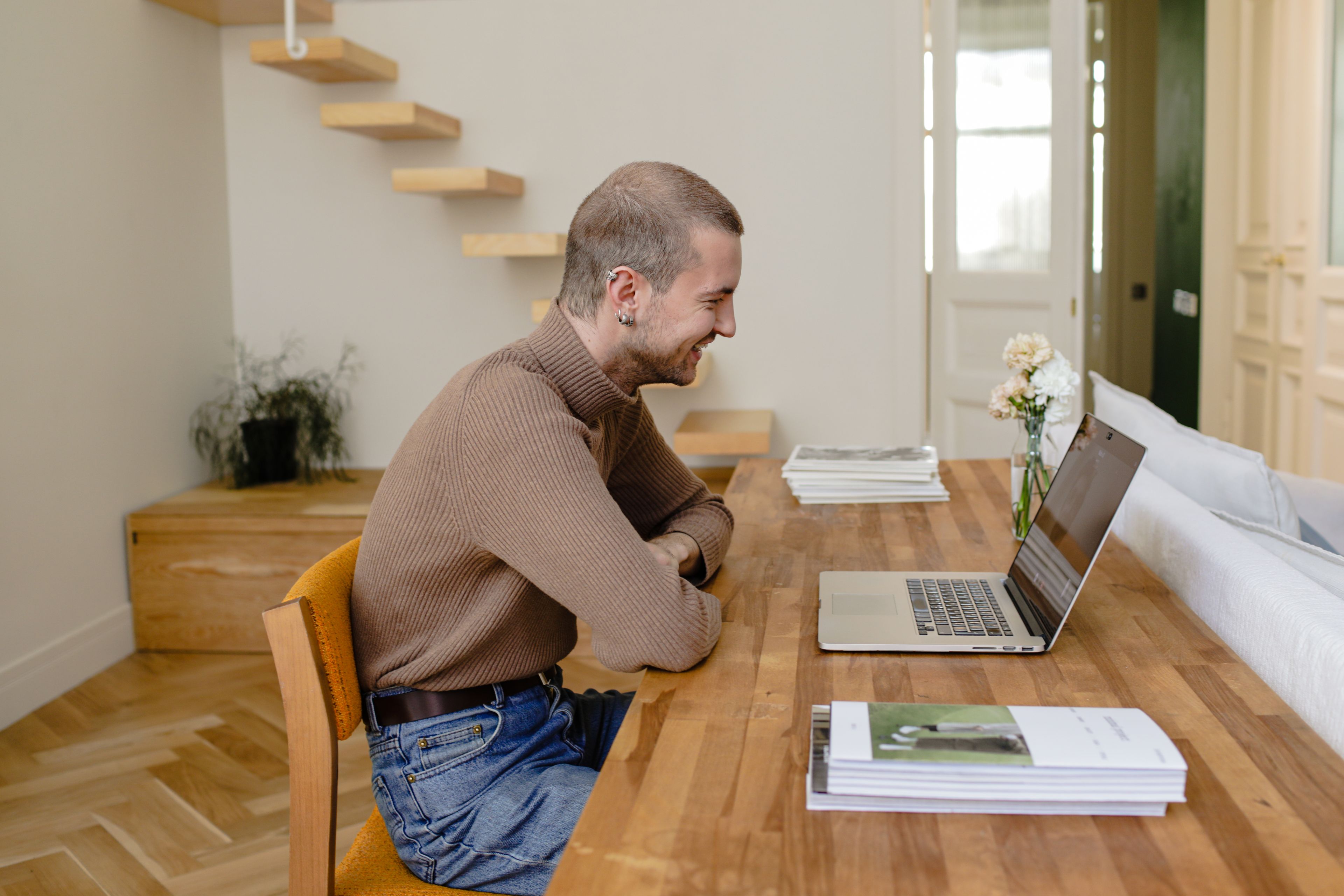 A man in a soft knit jumper talking with someone on a laptop video call at a warm wooden table in a calm, light-filled room