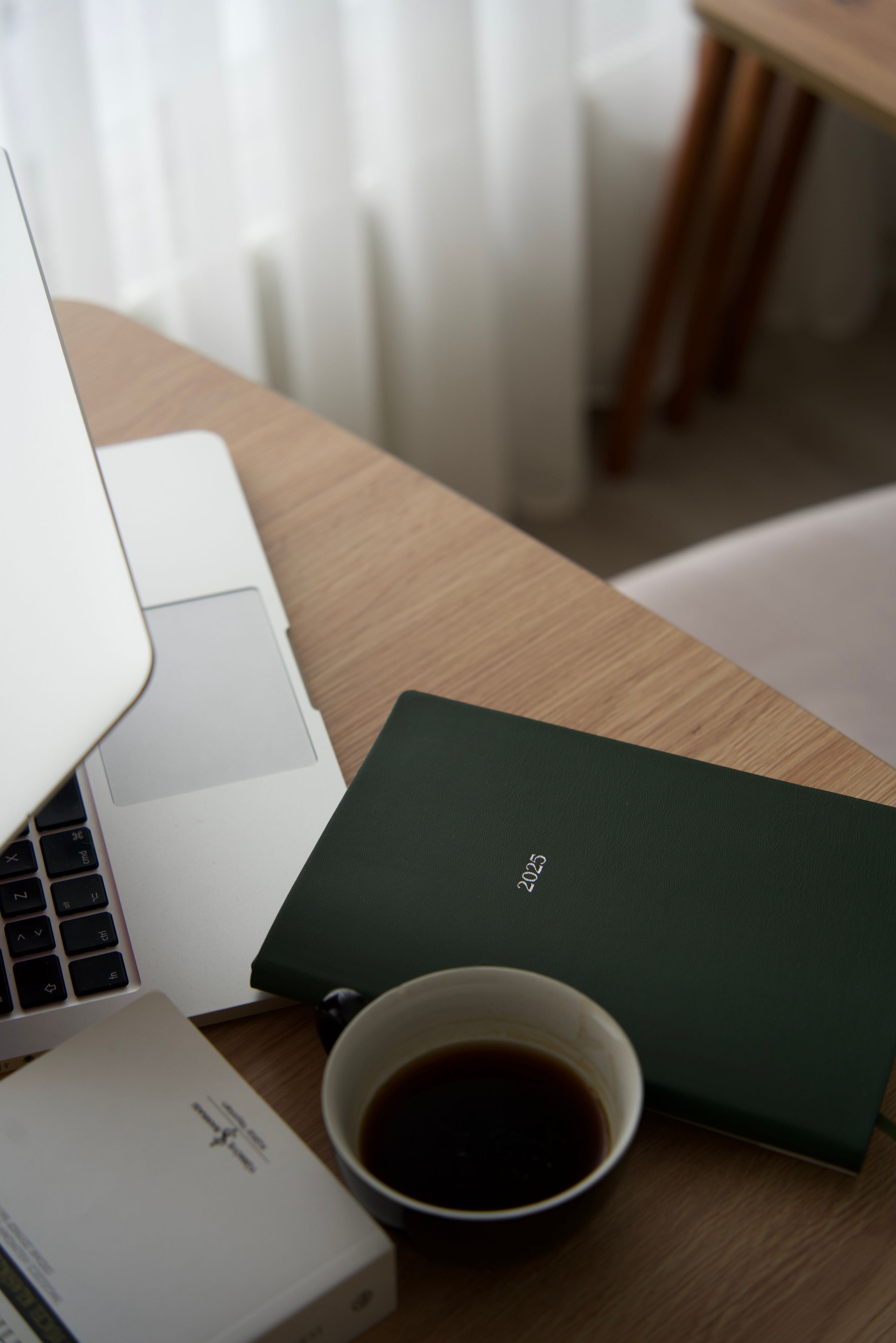 A laptop, a dark green leather planner and a cup of coffee on a warm timber desk in soft light