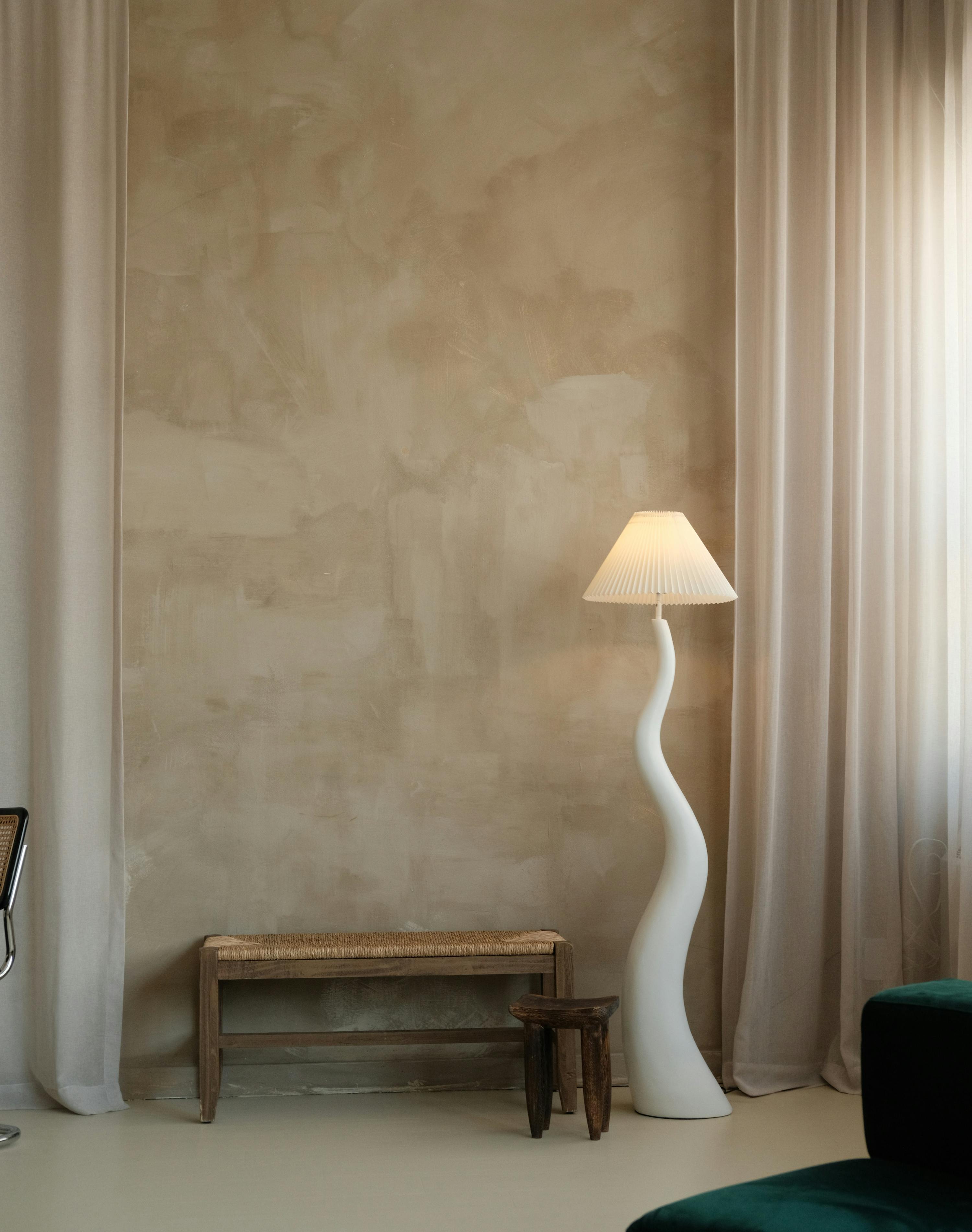 A calm, light-filled room with a sculptural lamp, a woven bench and soft linen curtains