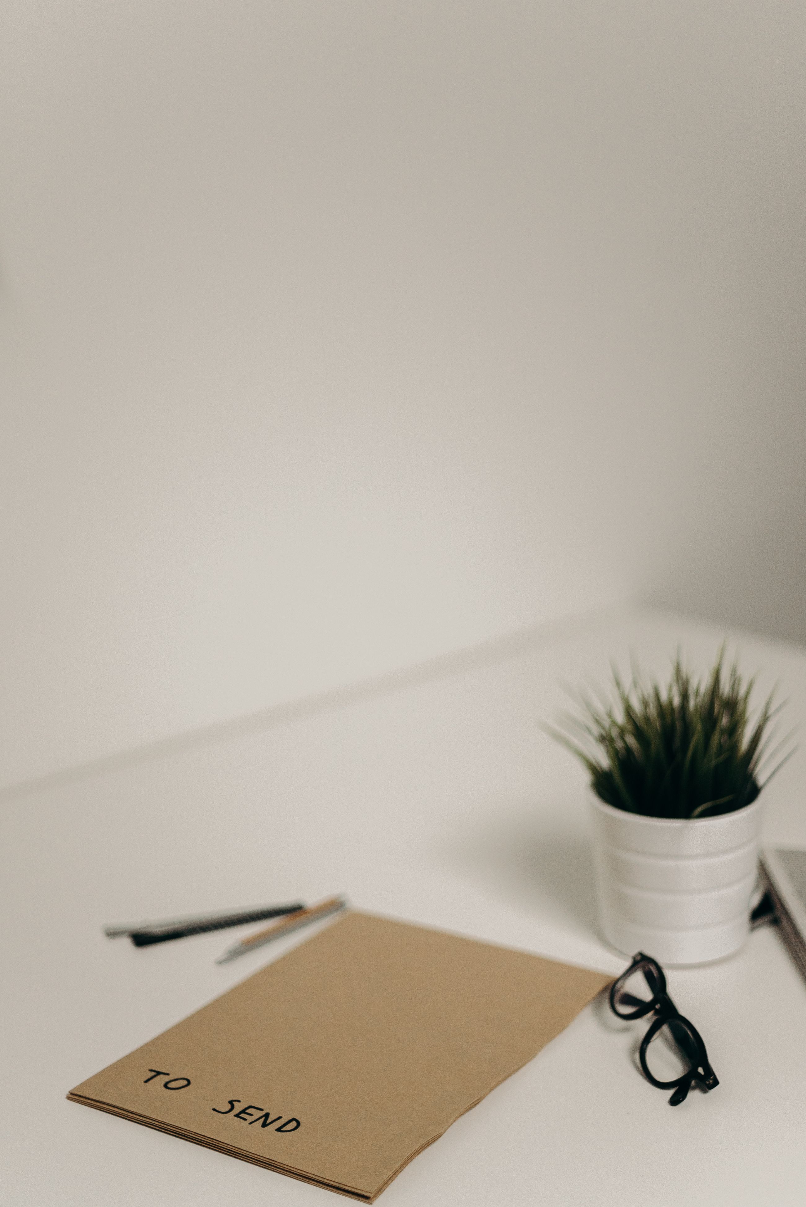 A kraft envelope marked to send, beside reading glasses, a pen and a small plant on a white desk