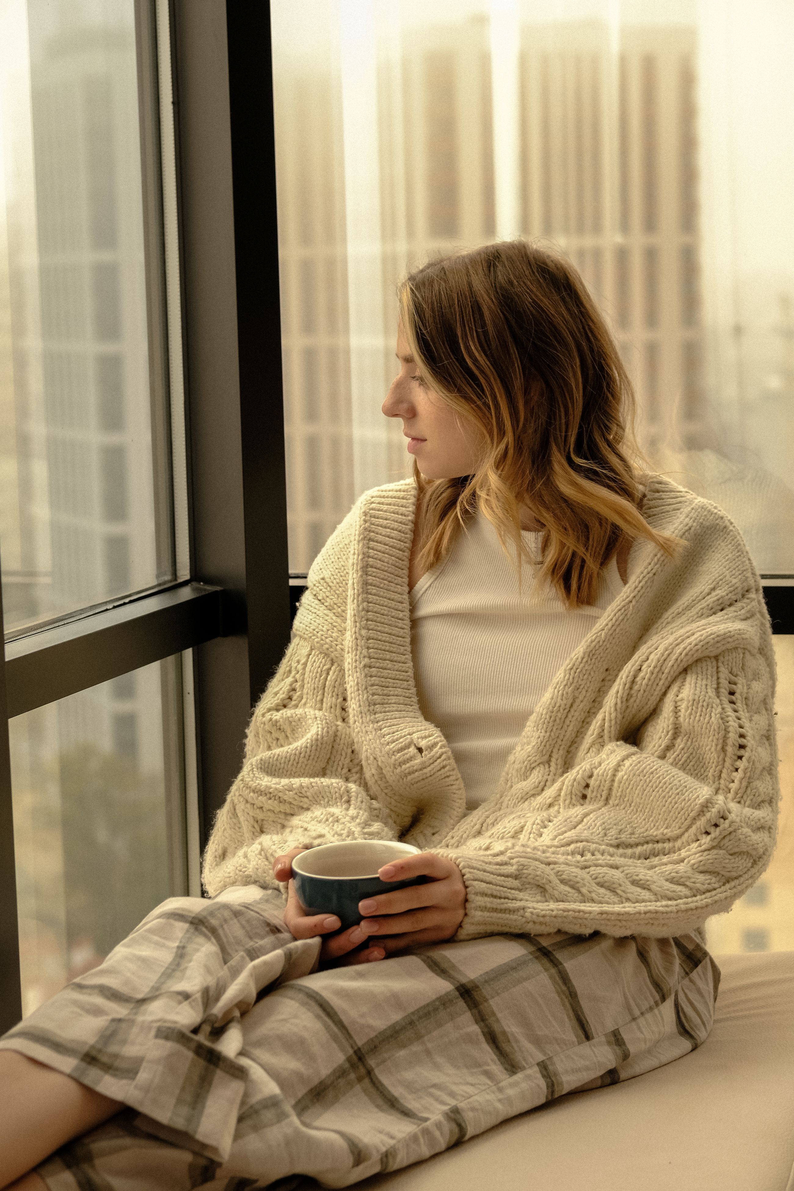A person in a soft knit cardigan sitting calmly by a window with a warm drink