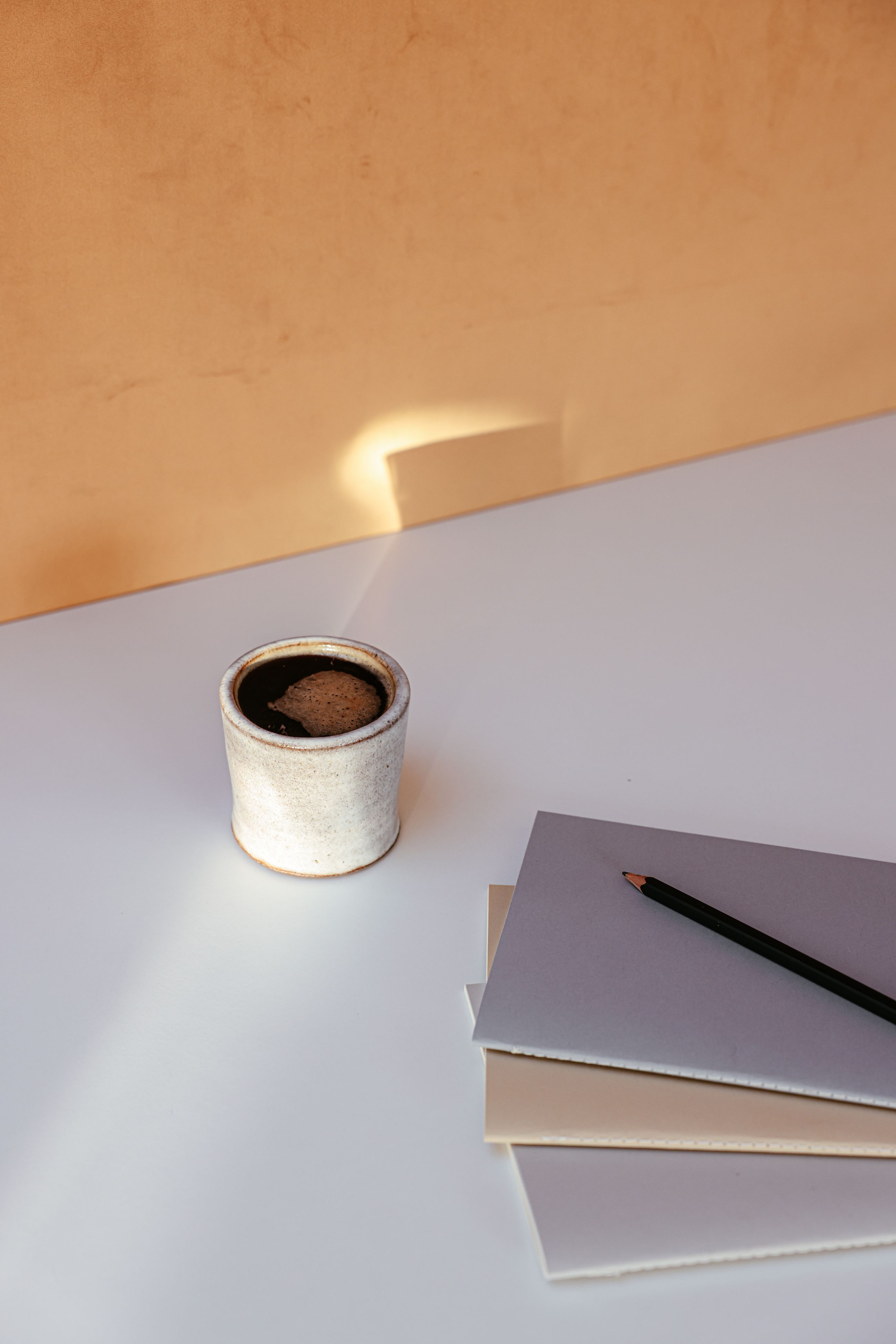A ceramic cup beside a neat stack of notebooks and a pencil on a desk in warm, quiet light