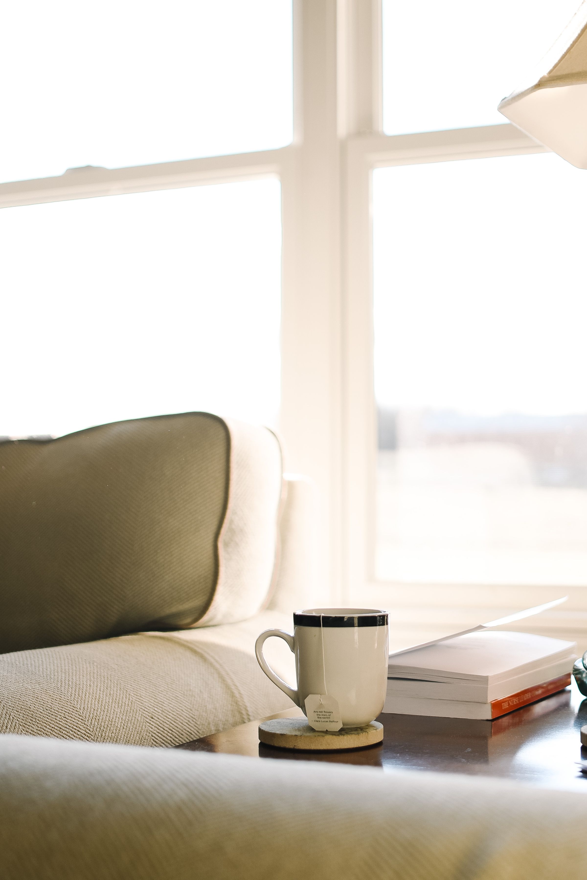 A warm mug and an open book on a side table beside a sofa, in a calm room with bright window light